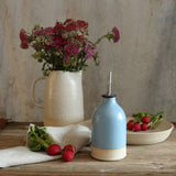 Small blue oil pourer on table next to a bowl of radishes and a jug of wildflowers