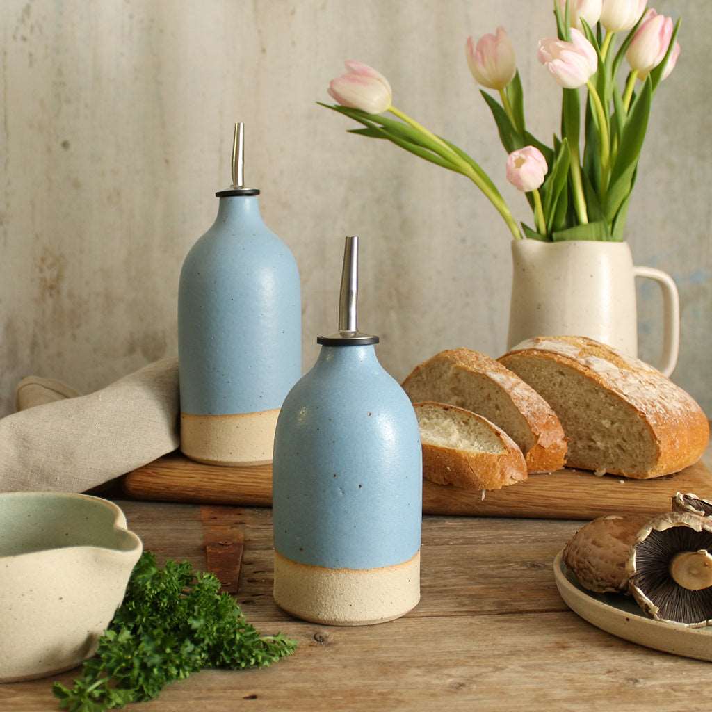 Blue small and large oil pourer on table surrounded by food with a jug of flowers in background