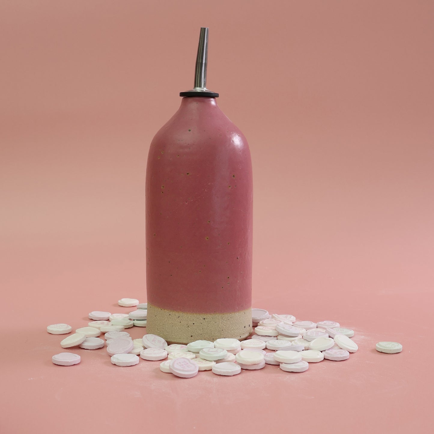 Bright pink handnmade stoneware oil pourer, surrounded by love heart sweets on a lighter pink background 