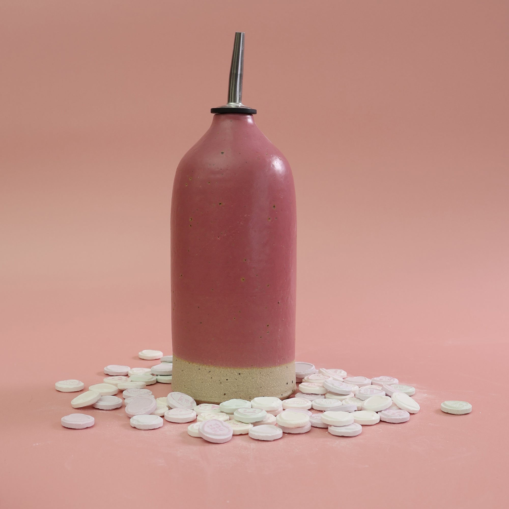 Bright pink handnmade stoneware oil pourer, surrounded by love heart sweets on a lighter pink background 
