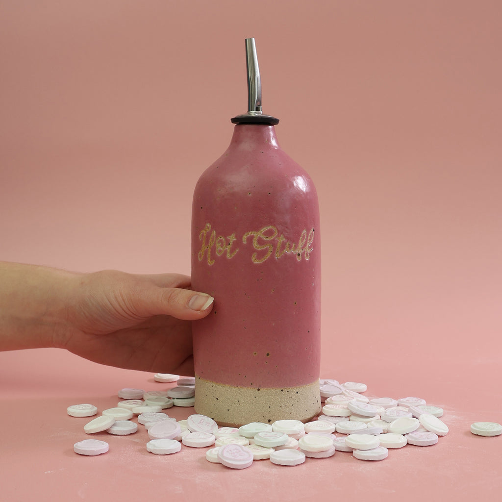 Pink oil pourer bottle with exposed clay lettering spelling 'Hot Stuff' with a pouring nozzle, surrounded by love heart sweets on a pink background