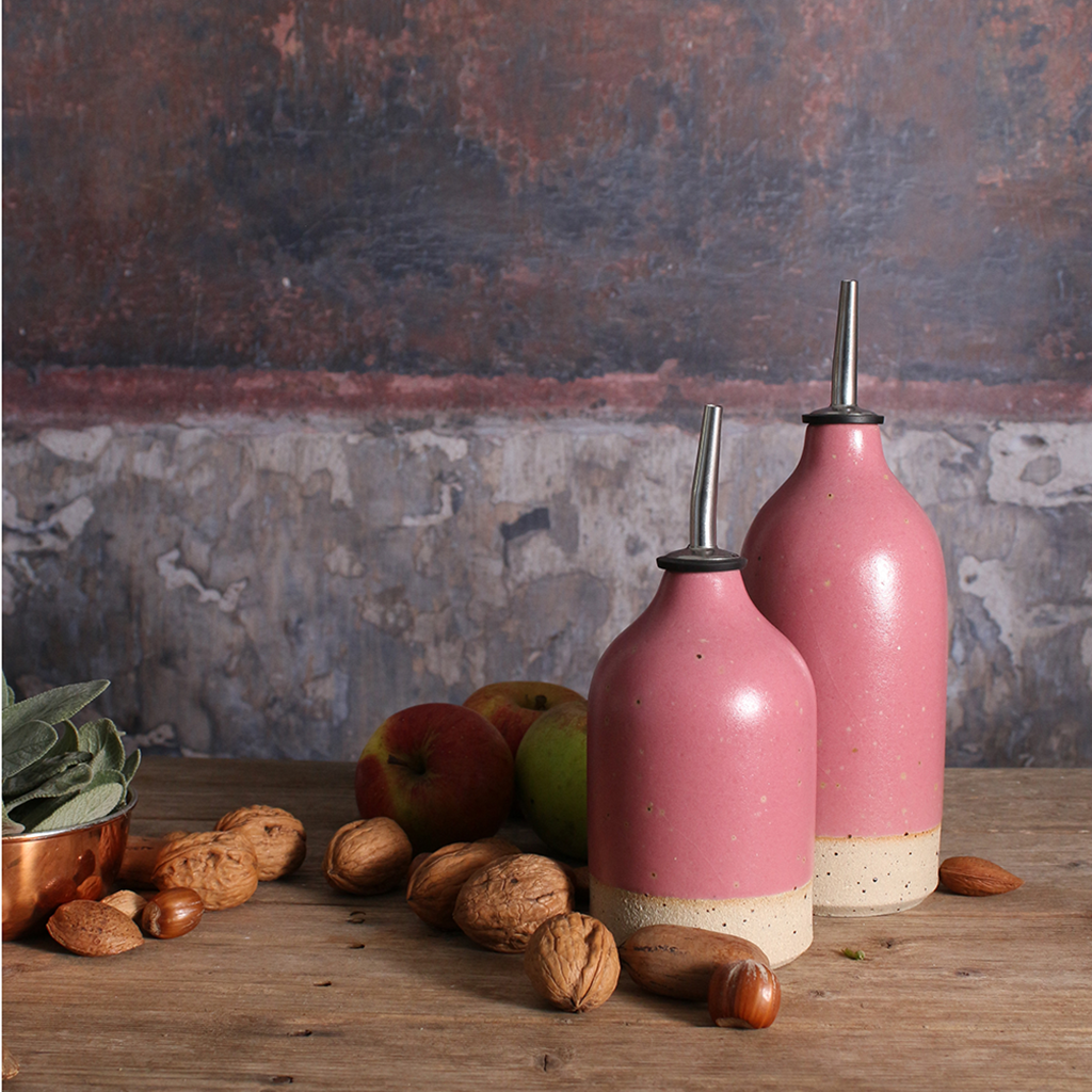 two fuchsia pink ceramic oil pourers in various sizes on a wooden table surrounded by walnuts and apples