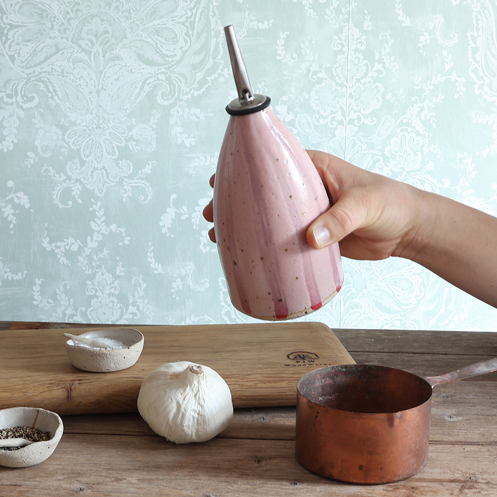 A hand holds a pink striped handmade ceramic oil dispenser with a stainless steel pour spout over a rustic wooden table with garlic, a copper pot, and small handmade bowls of ingredients.