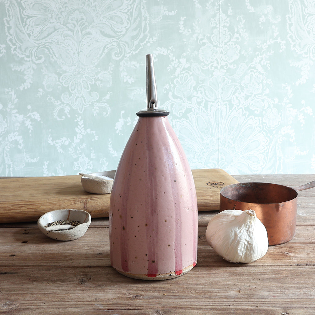 A pink striped handmade ceramic oil bottle with a stainless steel pour spout, styled on a rustic wooden table with a garlic bulb, a copper pot, and small bowls against a pale patterned wallpaper background.