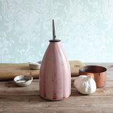 A pink striped handmade ceramic oil bottle with a stainless steel pour spout, styled on a rustic wooden table with a garlic bulb, a copper pot, and small bowls against a pale patterned wallpaper background.
