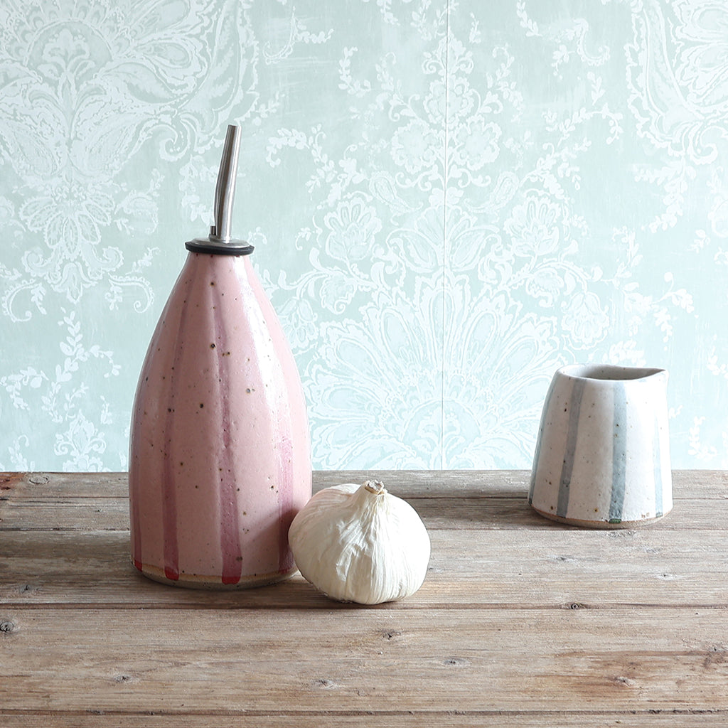 A pink striped handmade ceramic oil bottle with a stainless steel pour spout and a small white and blue stripe ceramic jug, styled on a rustic wooden table with a whole garlic bulb in front, against a pale wallpaper background.