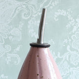 A close-up of a handmade ceramic oil pourer with a pink stripe design and a stainless steel pour spout, set against a pale green damask wallpaper background.