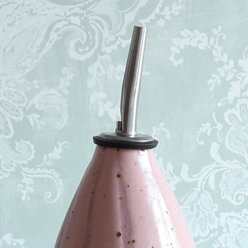 A close-up of a handmade ceramic oil pourer with a pink stripe design and a stainless steel pour spout, set against a pale green damask wallpaper background.
