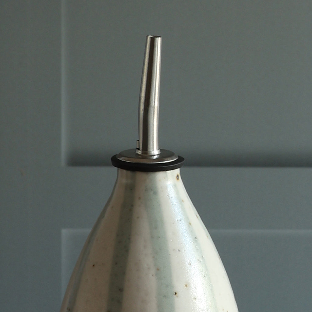 Close-up of a handmade ceramic oil pourer with a blue and white stripe design and stainless steel sillicone spout, set against a paneled background.