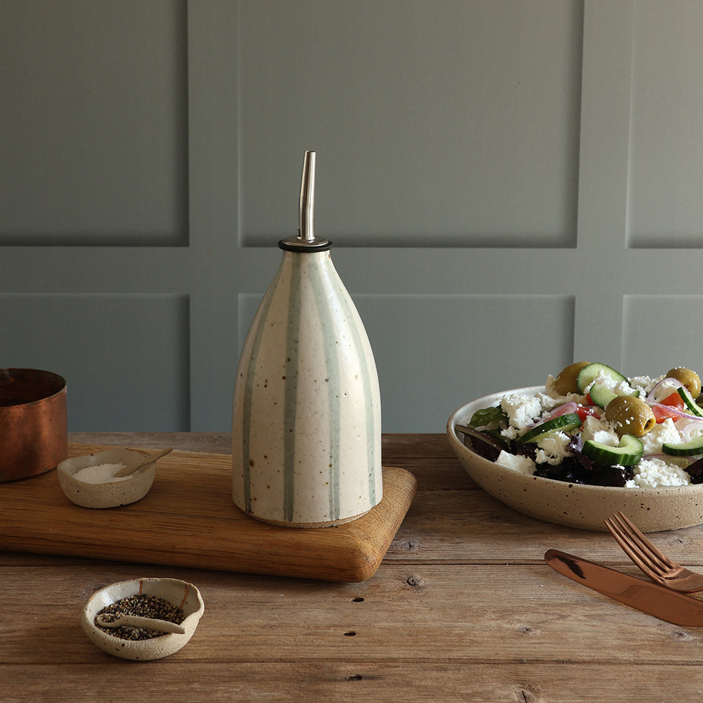 A handmade ceramic oil dispenser with a blue and white stripe design, and a stainless steel pour spout, styled on a wooden board on a rustic table next to a salad bowl.