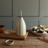 A handmade ceramic oil dispenser with a blue and white stripe design, and a stainless steel pour spout, styled on a wooden board on a rustic table next to a salad bowl.