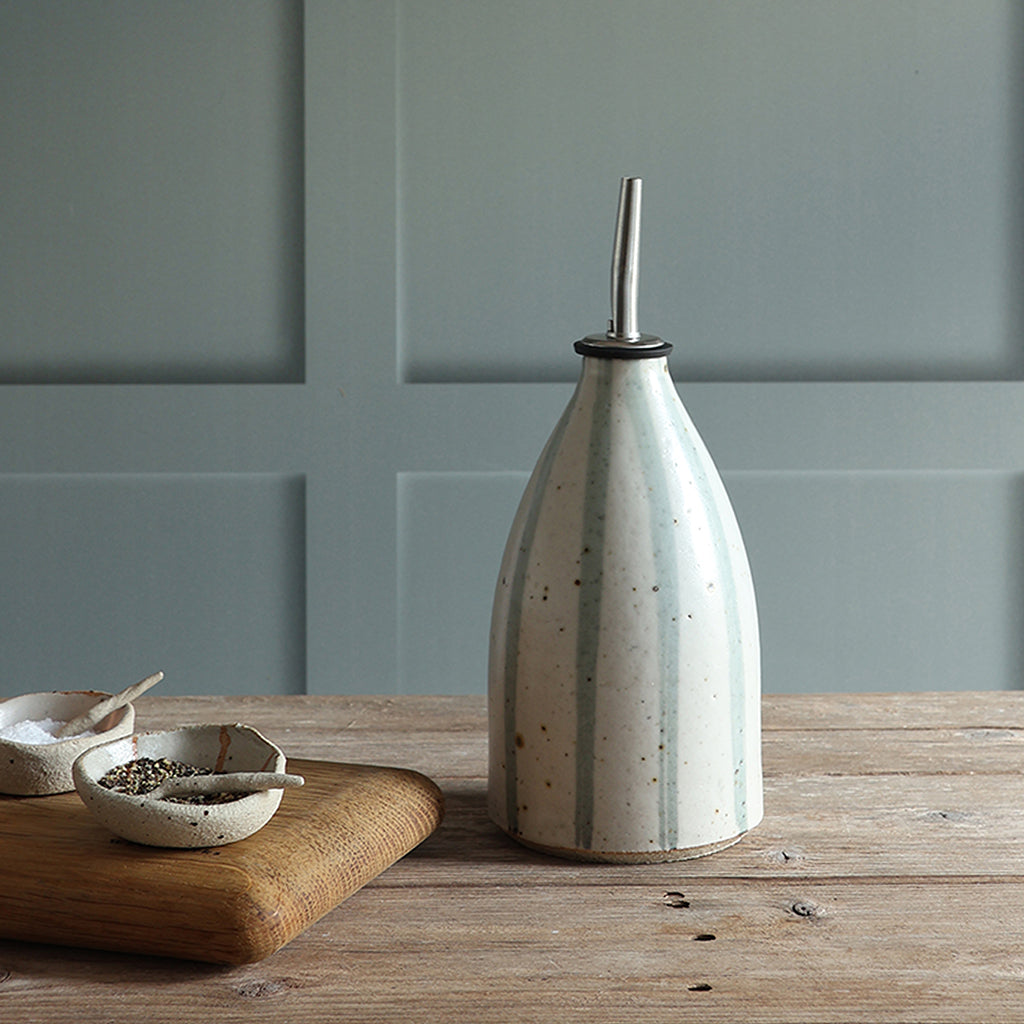 A ceramic oil pourer bottle with a stainless steel pour spout, featuring a speckled off-white glaze with vertical blue stripes, set on a wooden table against a panelled background. 