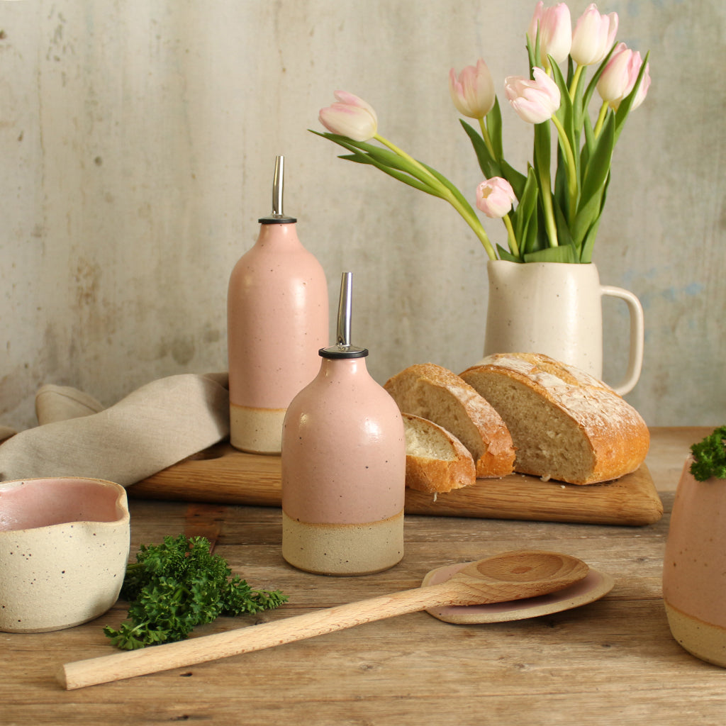 Pink small and large oil pourer on table with bread, wooden spoon, bowls and jug of flowers