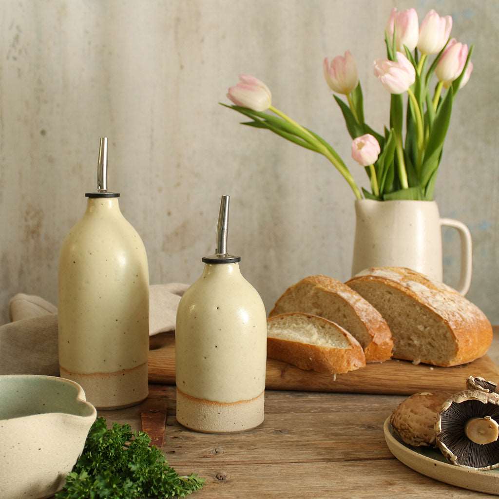 Yellow small and large oil pourer on table surrounded by food and a vase of flowers in background