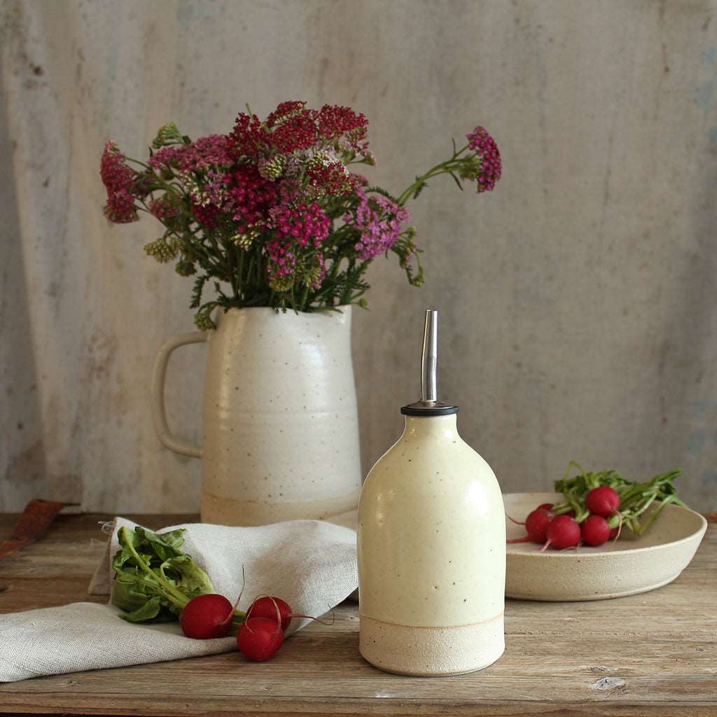 Small yellow oil pourer on table with bowl of radishes and jug of wildflowers