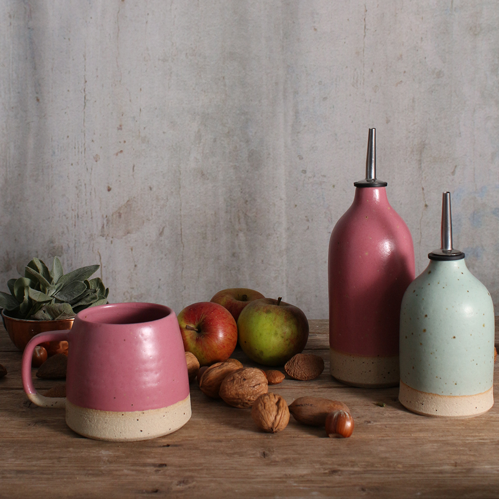 a fuchsia pink and duck egg blue ceramic oil pourers in various sizes on a wooden table surrounded by a fuchsia pink mug, walnuts and apples