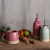 a fuchsia pink and duck egg blue ceramic oil pourers in various sizes on a wooden table surrounded by a fuchsia pink mug, walnuts and apples