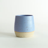 Front view of the cornflower blue glazed tumbler with exposed raw flecked clay around the base. Dark flecks from the clay can be seen through the glaze.