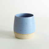 A cornflower blue glazed tumbler with exposed raw flecked clay around the base. Dark flecks from the clay can be seen through the glaze.