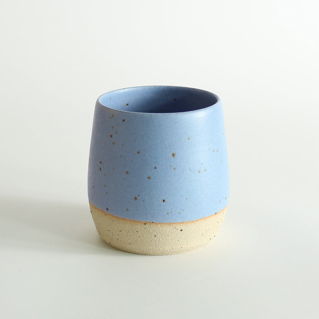 A cornflower blue glazed tumbler with exposed raw flecked clay around the base. Dark flecks from the clay can be seen through the glaze.