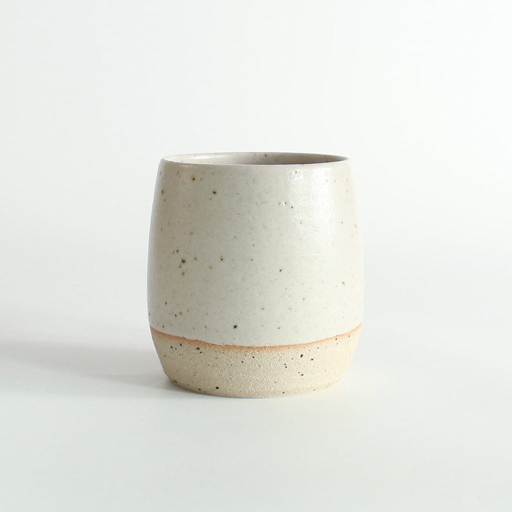 Front view of the off-white glazed tumbler with exposed raw flecked clay around the base. Dark flecks from the clay can be seen through the glaze.