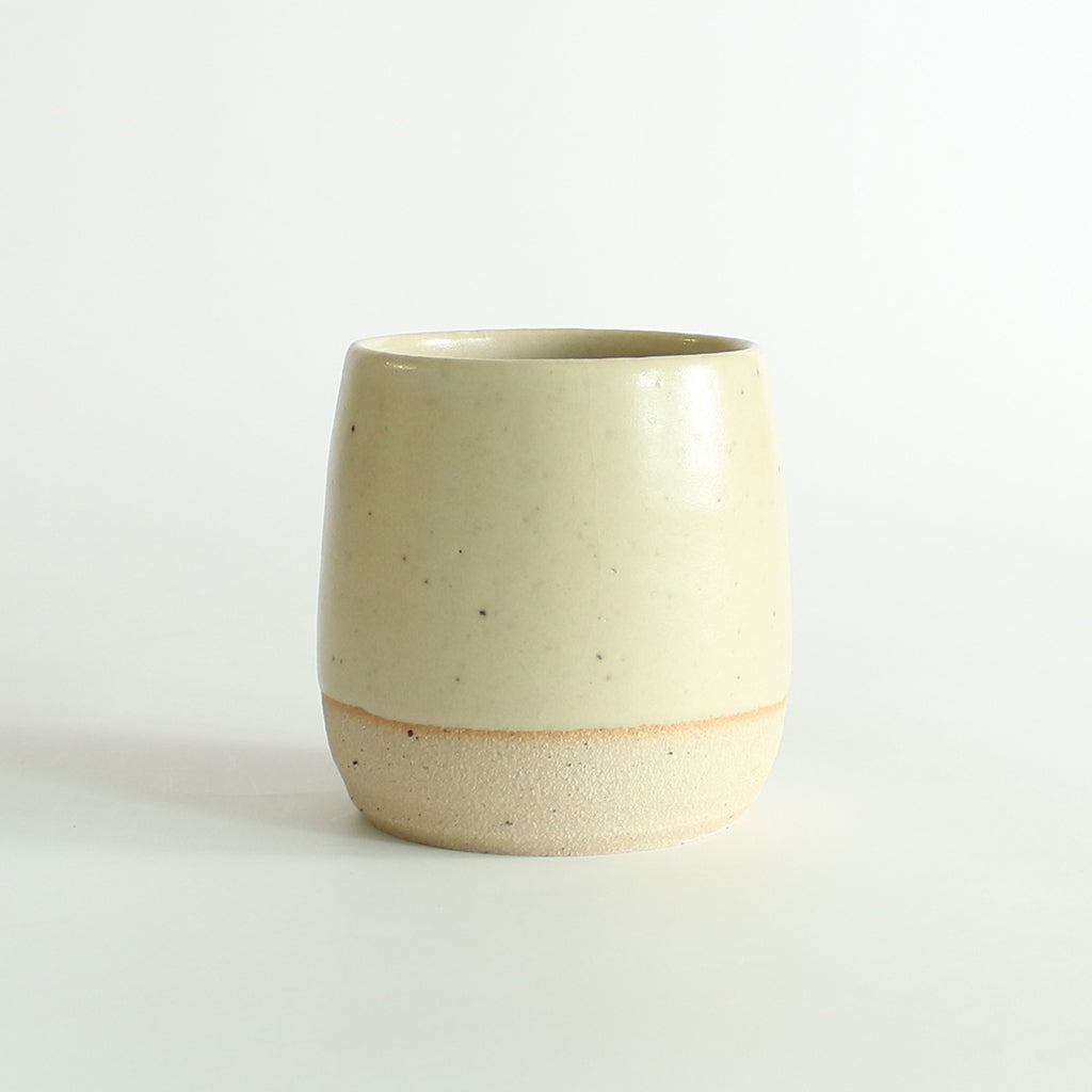 Front view of the light yellow glazed tumbler with exposed raw flecked clay around the base. Dark flecks from the clay can be seen through the glaze.