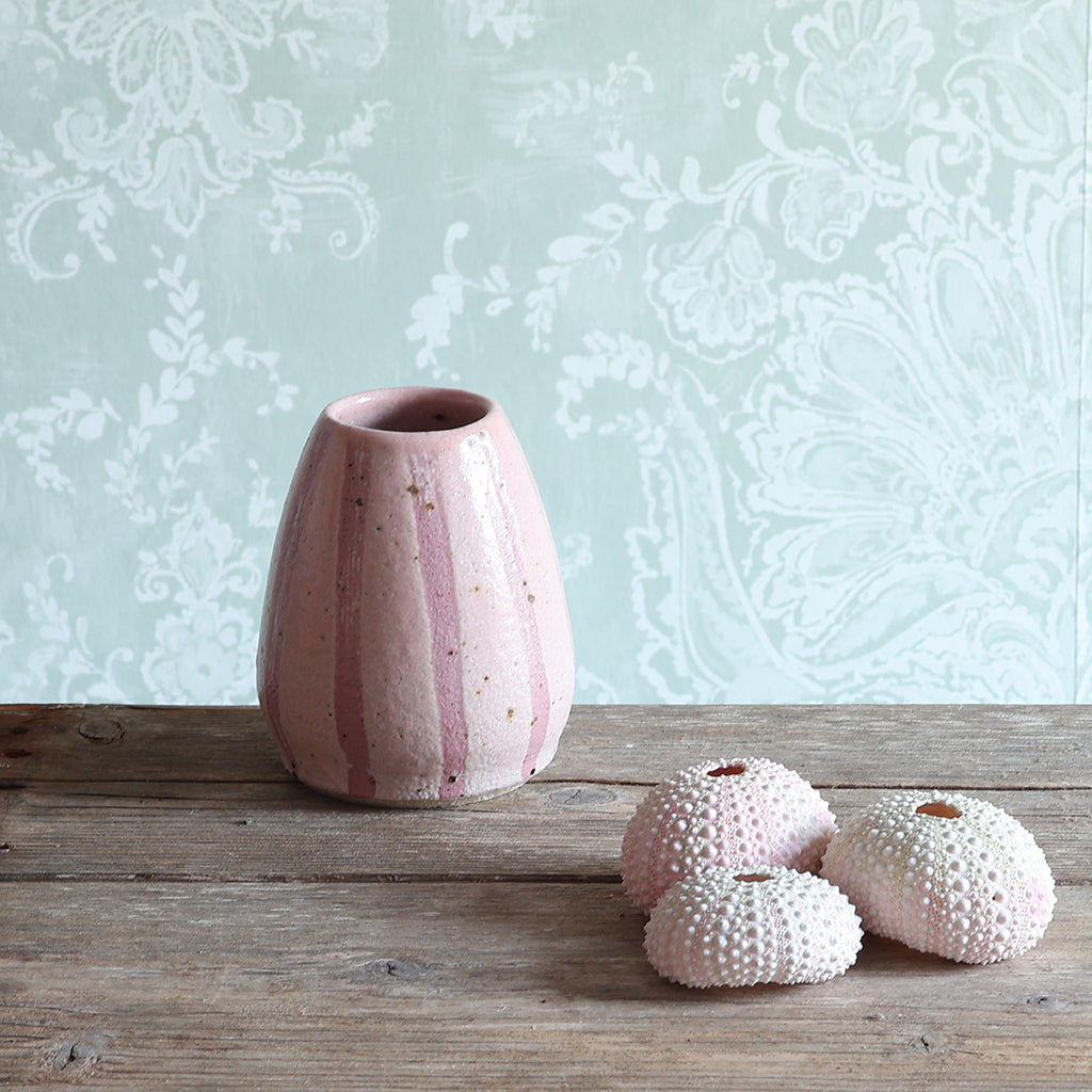 A pink ceramic vase with vertical pink stripes sits on a rustic wooden table next to a collection of three sea urchin shells. 