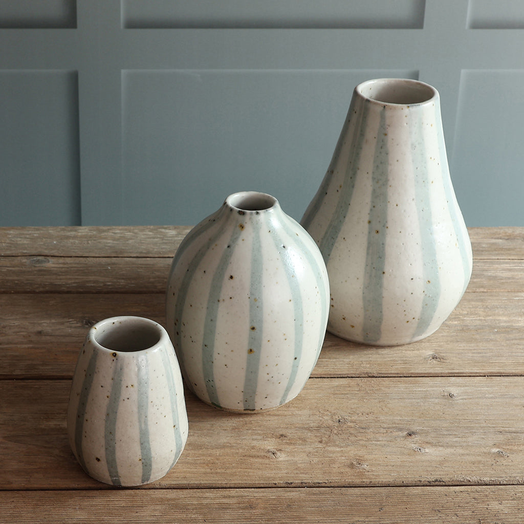 Three handmade ceramic vases with vertical blue and white stripes and, displayed on a rustic wooden table against a blue paneled wall.