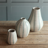 Three handmade ceramic vases with vertical blue and white stripes and, displayed on a rustic wooden table against a blue paneled wall.