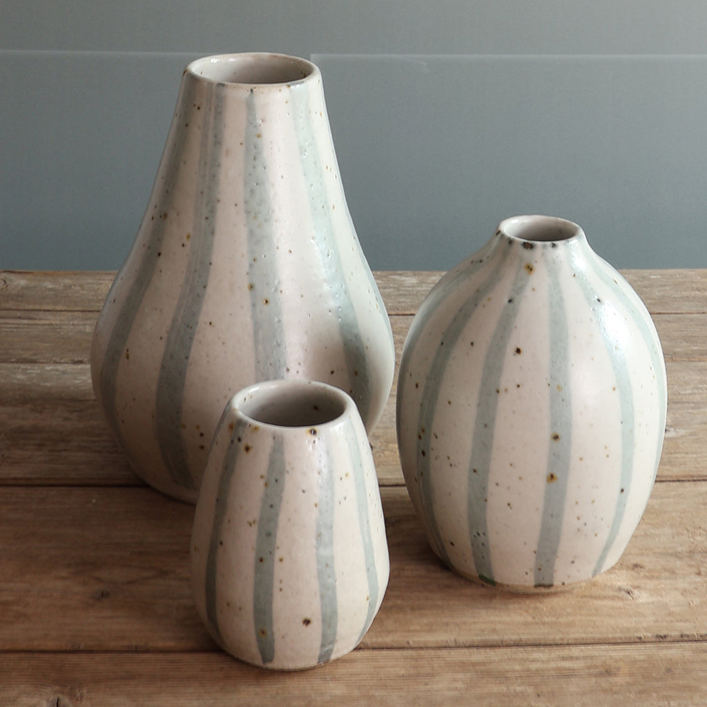 Three handmade ceramic vases in varying heights with vertical blue and white stripes on a rustic wooden table against a panelled wall.