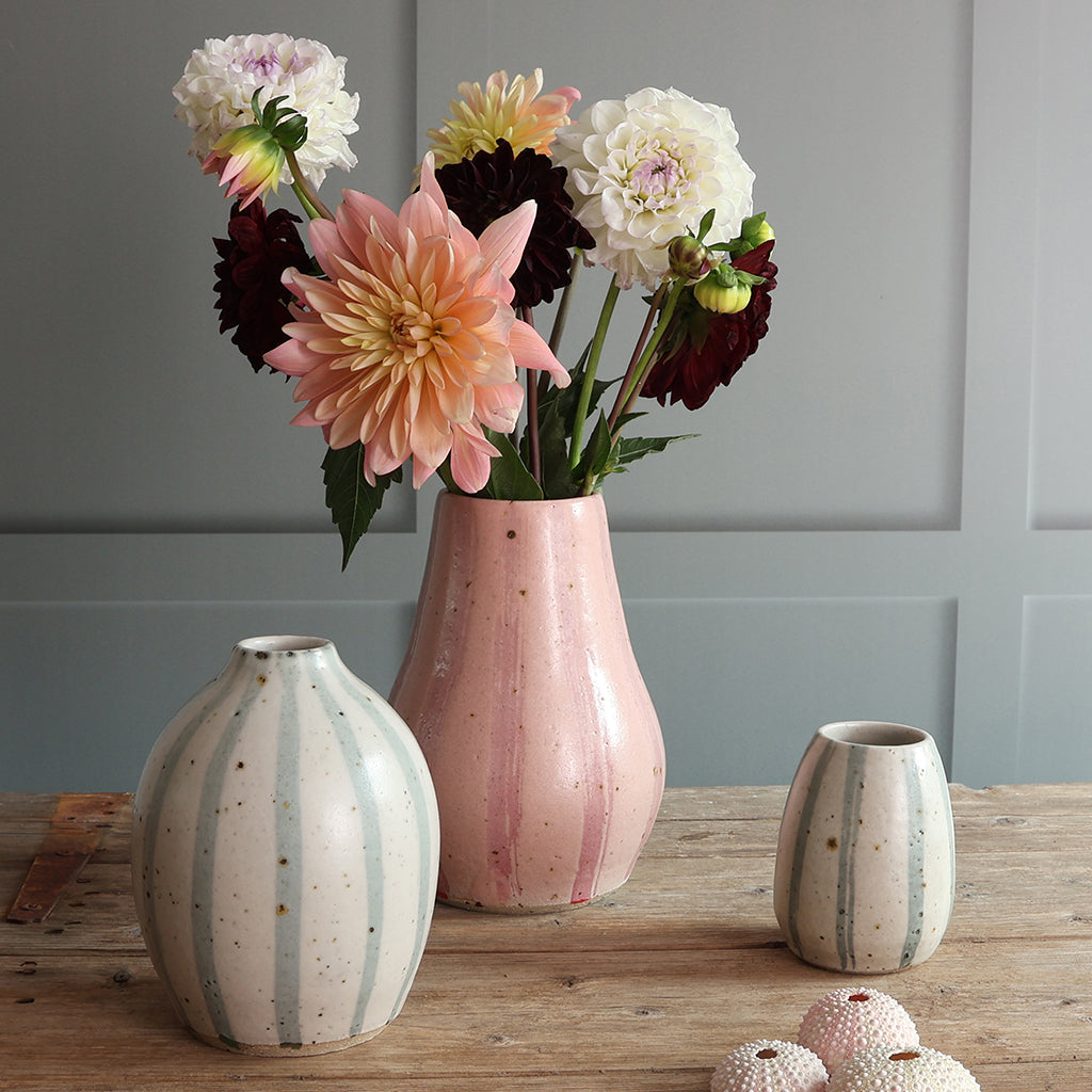 Three ceramic vases in a styled tabletop arrangement, with a pink stripey vase holding a bouquet of dahlias against a gray paneled wall. The other vases feature a blue and white stripe design. 
