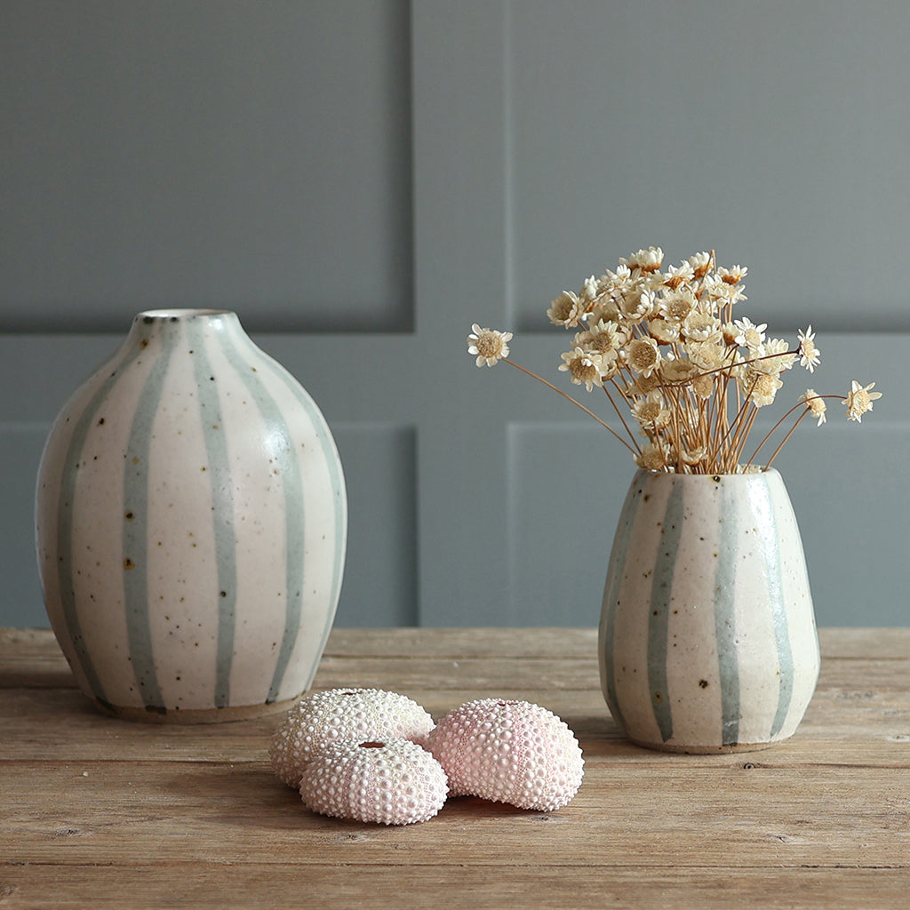 Two handmade ceramic vases with blue and white stripes, displayed on a wooden surface against a paneled wall. One vase is empty, and the other holds dried flowers.