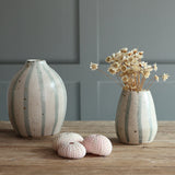 Two handmade ceramic vases with blue and white stripes, displayed on a wooden surface against a paneled wall. One vase is empty, and the other holds dried flowers.