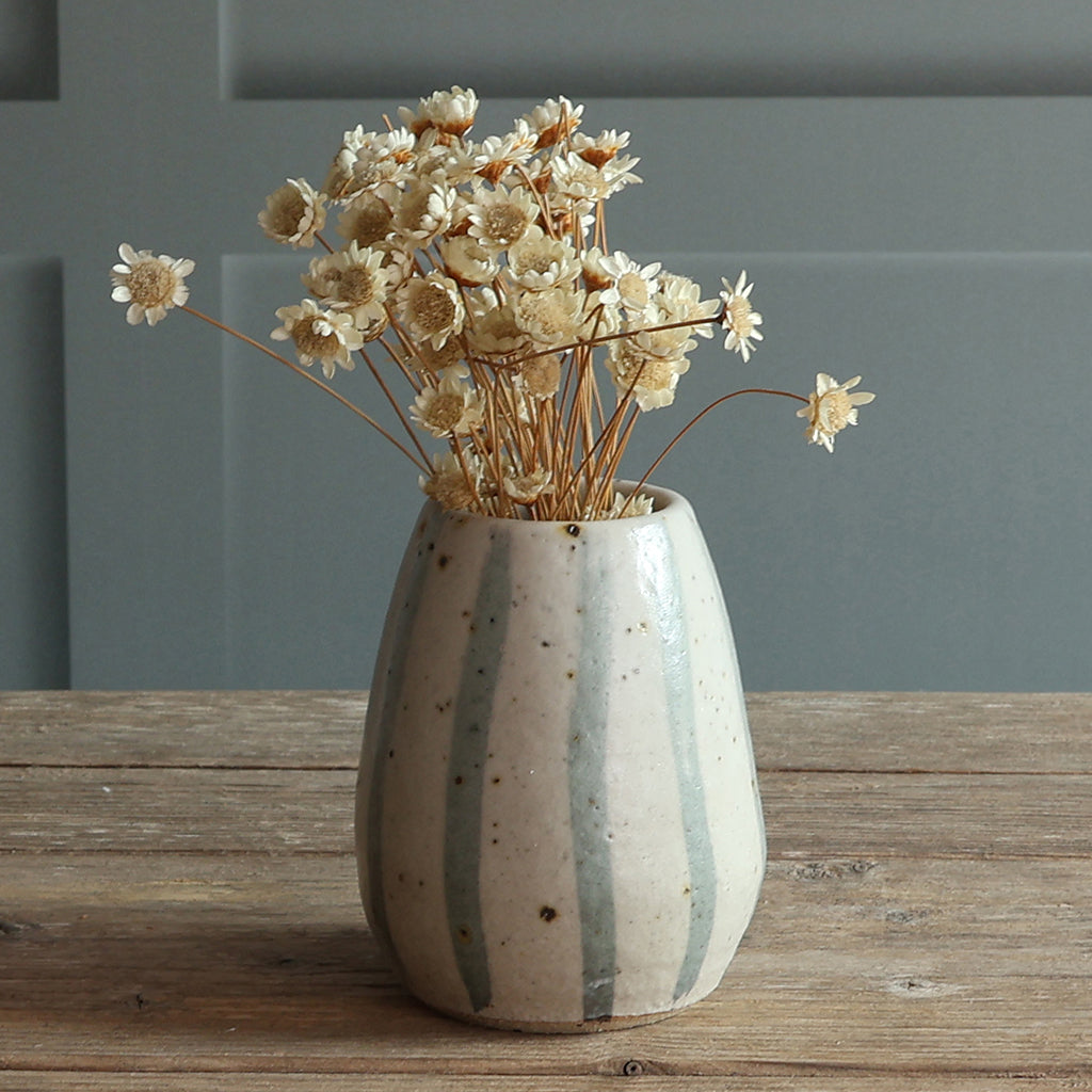 A small speckled ceramic vase with blue and white stripes, filled with cream flowers, set on a rustic wooden tabletop against a paneled wall.