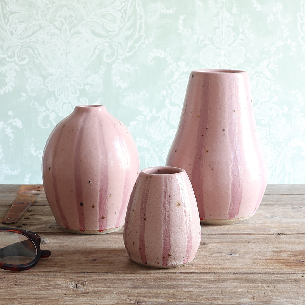 Three pink ceramic vases of different sizes with vertical pink stripes, arranged on a rustic wooden table against a pale green damask wallpaper background. 
