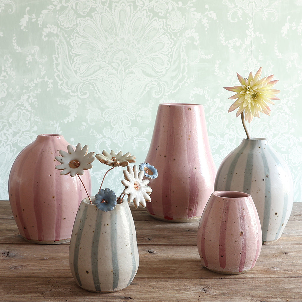 Five handmade ceramic vases in either a pink stripe or blue and white stripe design, arranged on a wooden surface against a pale green damask-patterned wall.