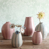 Five handmade ceramic vases in either a pink stripe or blue and white stripe design, arranged on a wooden surface against a pale green damask-patterned wall. One vase is filled with handmade ceramic flowers. 