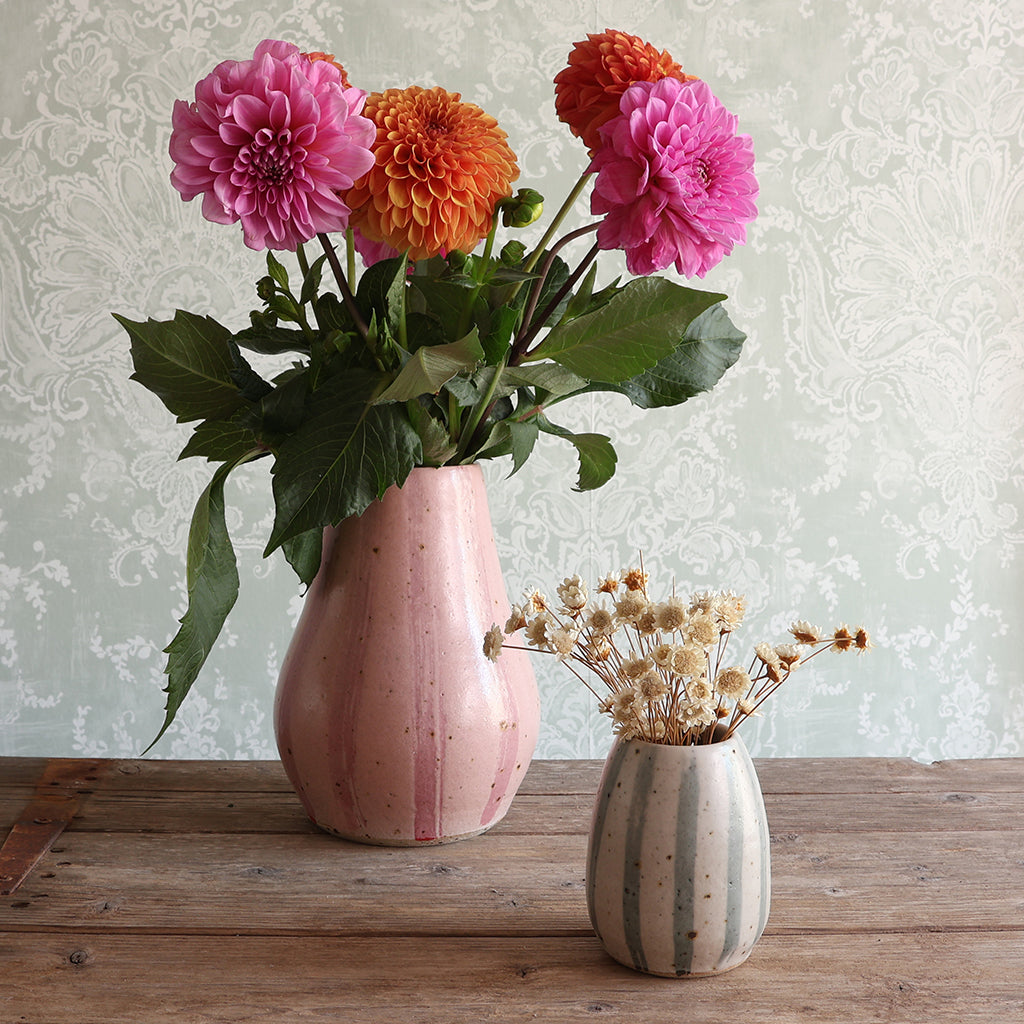 A pink striped ceramic vase filled with bright pink and orange dahlias and green leaves, placed on a rustic wooden table next to a smaller striped ceramic vase with dried flowers. 