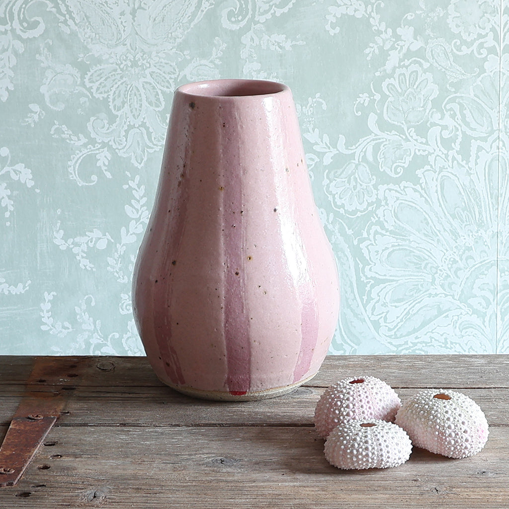 A handmade ceramic vase with vertical pink stripes, displayed on a rustic wooden surface next to three pink sea urchin shells against a pale green damask wallpaper background. 