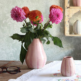 A pink handmade ceramic vase filled with bright pink and orange flowers sits on a wooden table, with a matching empty pink vase beside it and a patterned wallpapered room in the background.