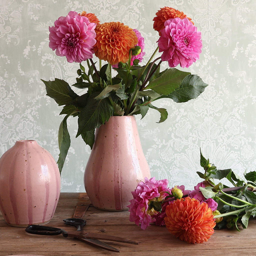 A handmade pink stripey ceramic vase filled with bright pink and orange dahlias sits on a rustic wooden table, alongside a matching pink vase, scissors, and additional flowers. 
