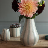 A ceramic vase with vertical blue and white stripes stands on a wooden table holding a peach dahlia flower, with a smaller matching vase and planter behind it and a panelled wall in the background.