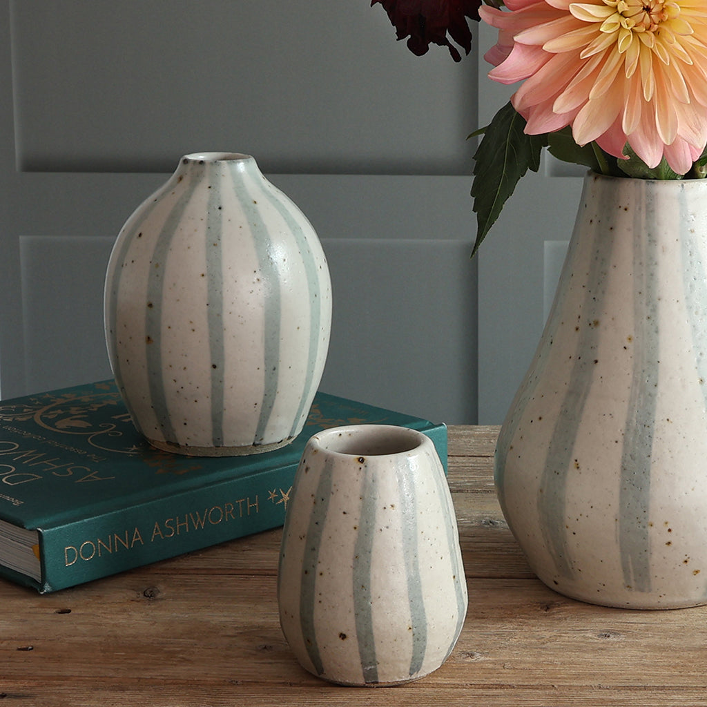 Three handmade ceramic vases with blue and white stripe designs, styled on a wooden table with a teal book. The largest vase holds a large pink and yellow flower. 