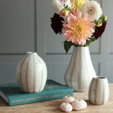 Three speckled ceramic vases with white and pale blue stripes arranged on a wooden table, with one vase holding a bouquet of dahlias. A teal book titled 'Donna Ashworth Joy Chess You' sits beneath the left vase.