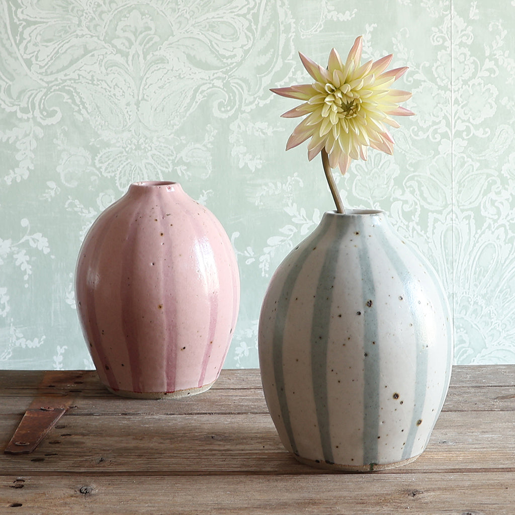 Two ceramic vases on a rustic wooden table. The vase on the left has a pink stripe design, and the vase on the right has a blue and white stripe design, and holds a pale yellow dahlia flower.