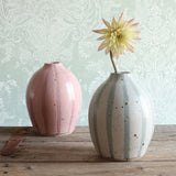 Two ceramic vases on a rustic wooden table. The vase on the left has a pink stripe design, and the vase on the right has a blue and white stripe design, and holds a pale yellow dahlia flower.