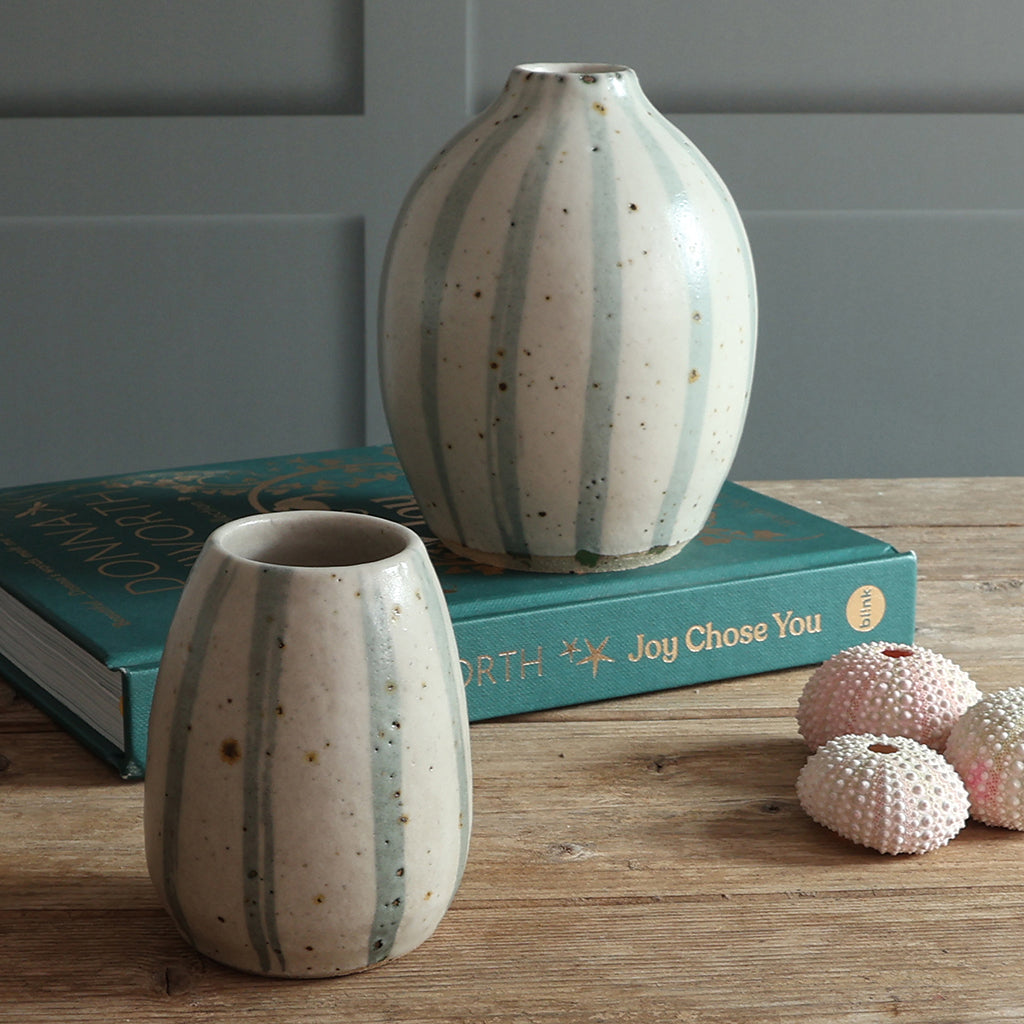 Two speckled ceramic vases with blue and white striped designs, styled on a wooden table with a teal book and pink shells against a paneled wall.