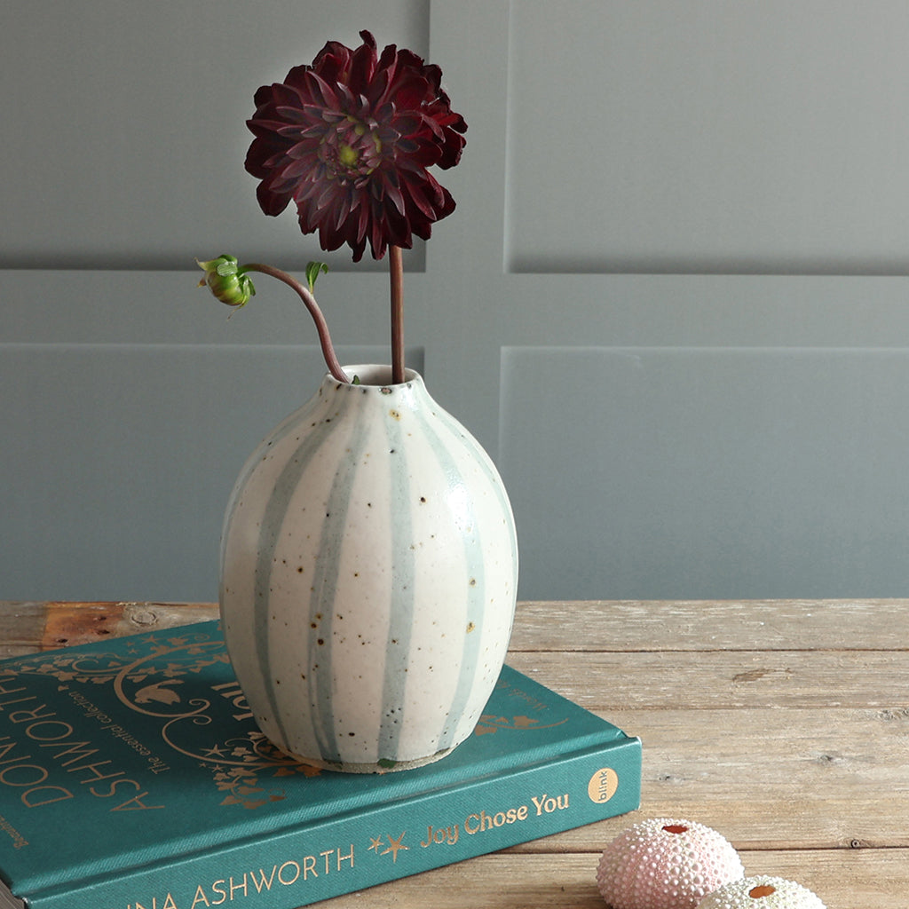 A handmade ceramic vase with vertical blue and white stripes sits on a teal book on a wooden table, holding a dark burgundy dahlia flower against a paneled wall.