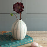 A handmade ceramic vase with vertical blue and white stripes sits on a teal book on a wooden table, holding a dark burgundy dahlia flower against a paneled wall.