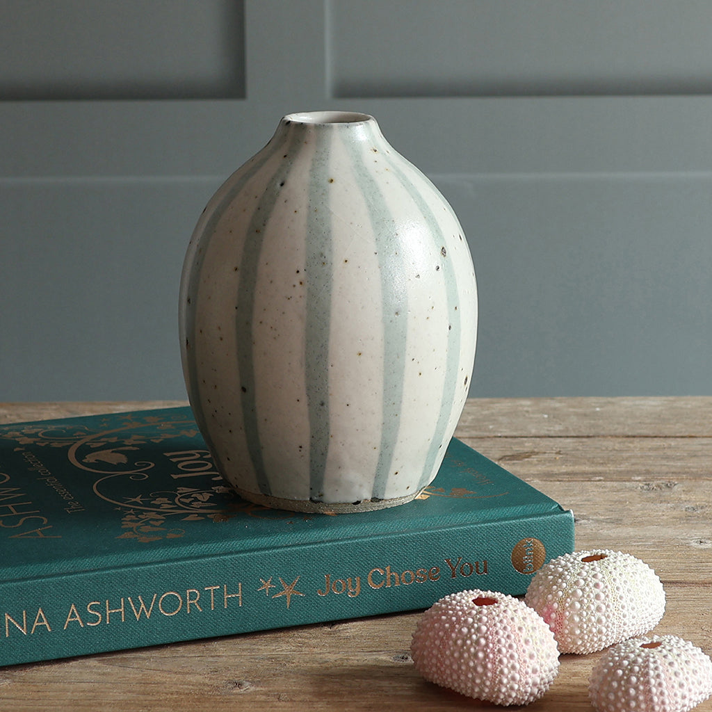 A handmade ceramic vase with vertical blue and white stripes, displayed on a teal book on a wooden surface with pink decorative shells in the foreground.